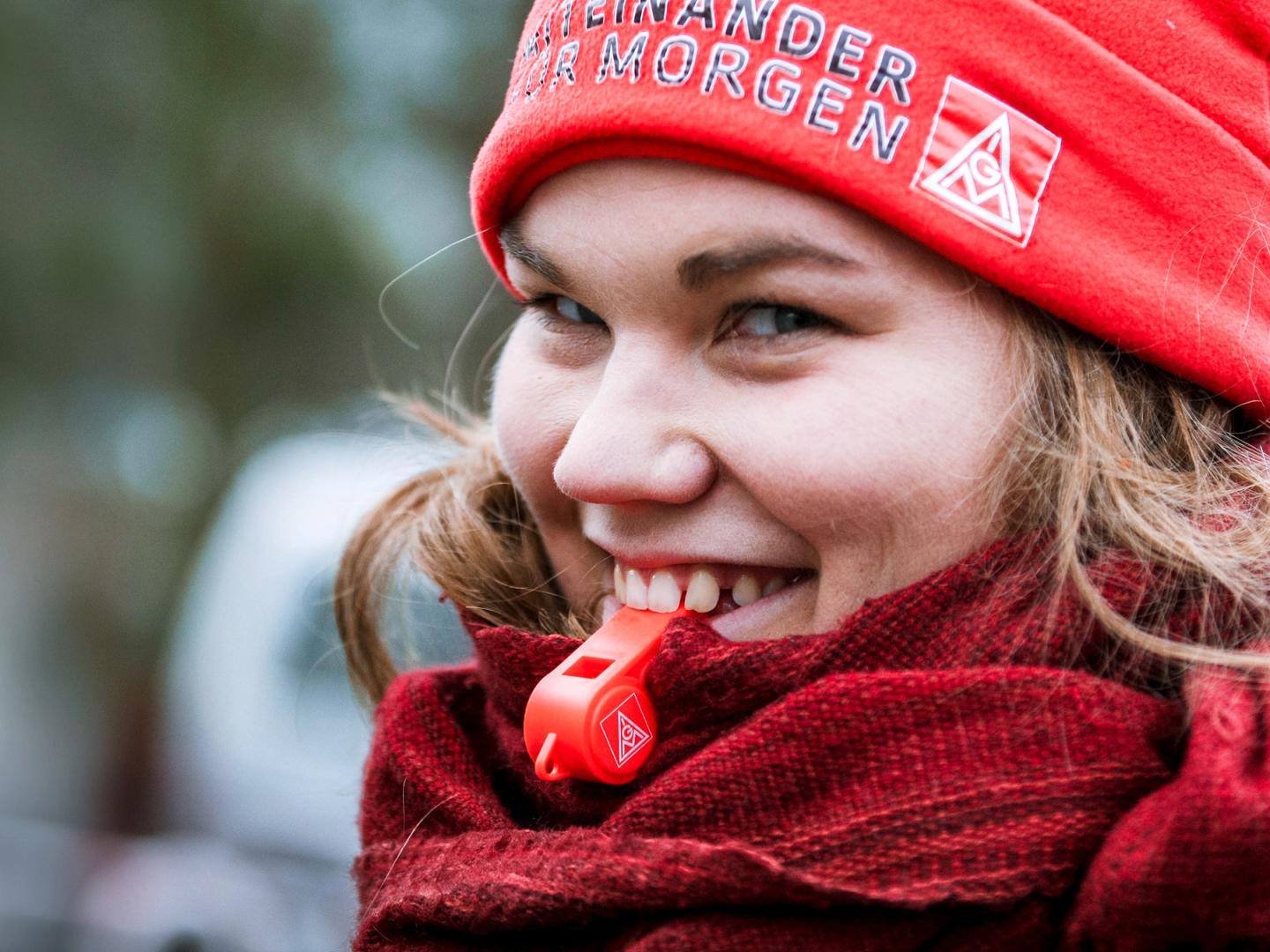Eine junge Frau mit blonden Haaren und einer roten IG Metall-Mütze hat eine rote Pfeife im Mund und lächelt.