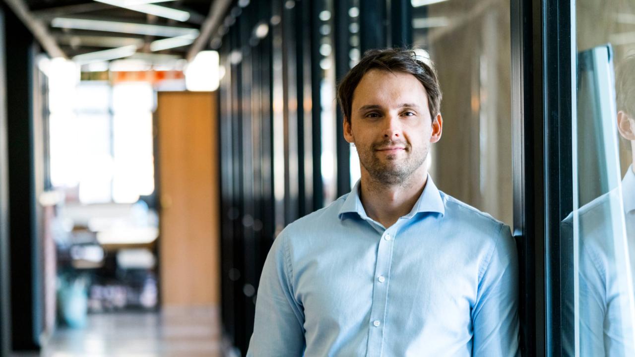 Ein lächelnder Mann mit blauem Hemd lehnt an einer Wand.