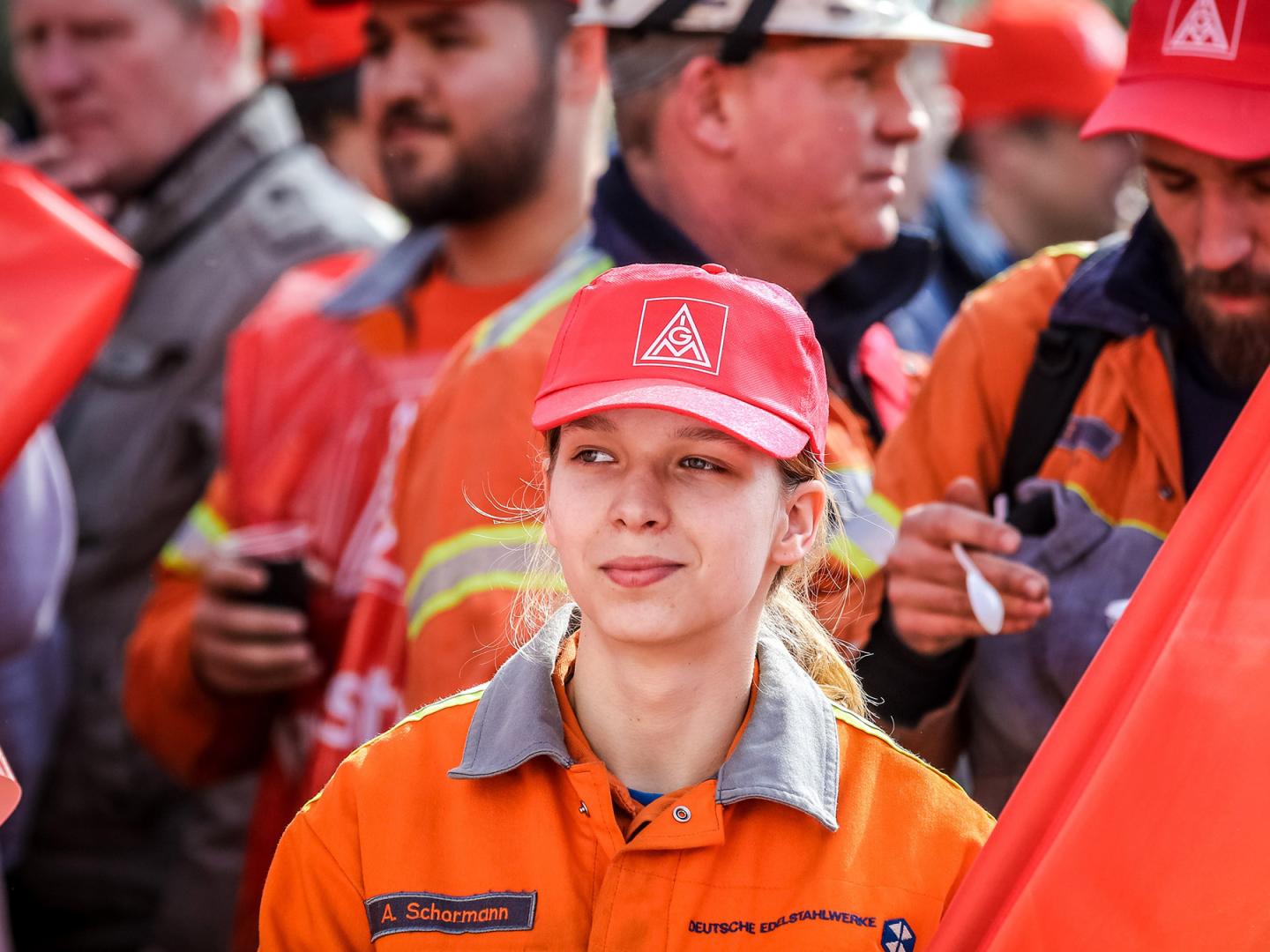 Eine junge Frau mit IG Metall-Kappe, einer Fahne und Arbeitskleidung auf einer Demo.