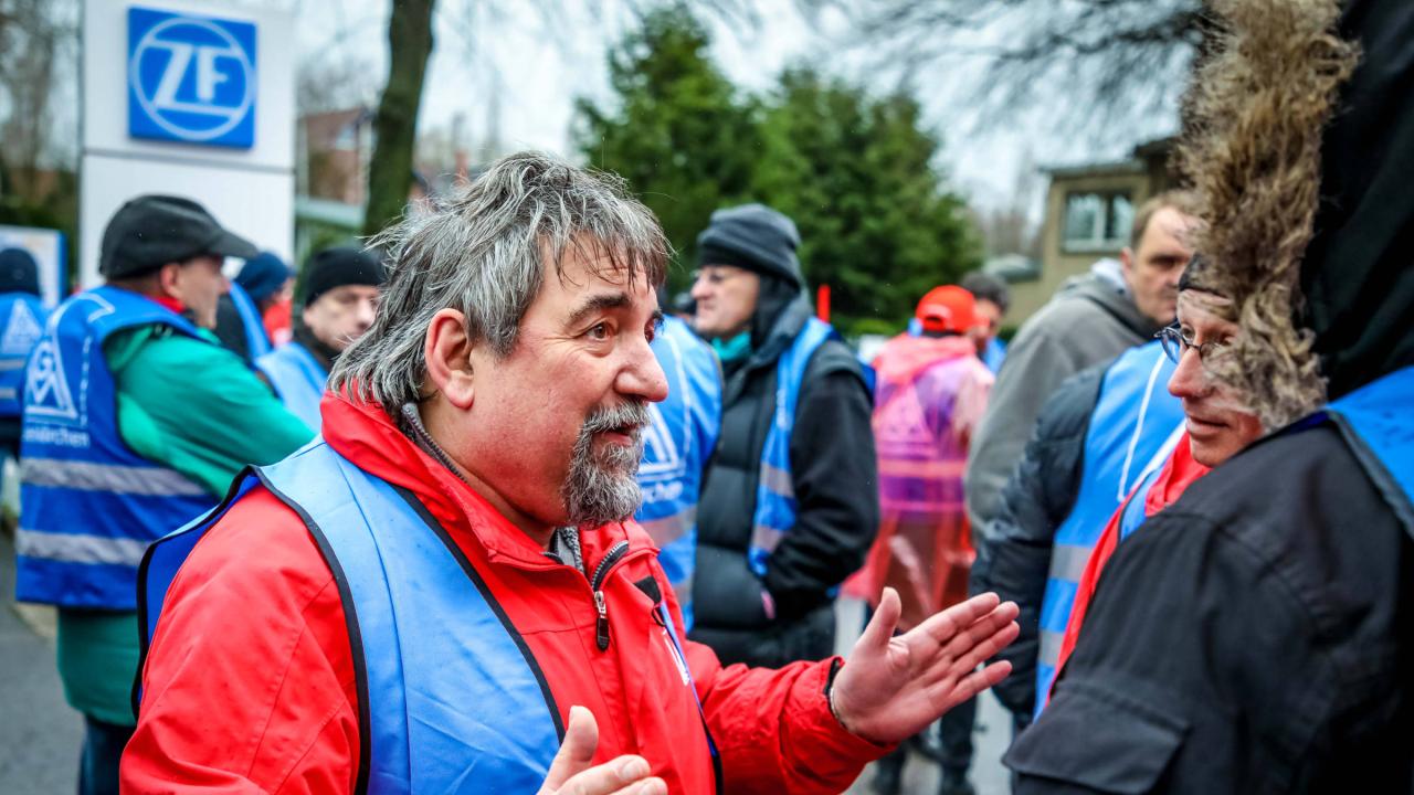 Ein Mann in einer blauen Streikeste im Gespräch mit einem anderen Mann.