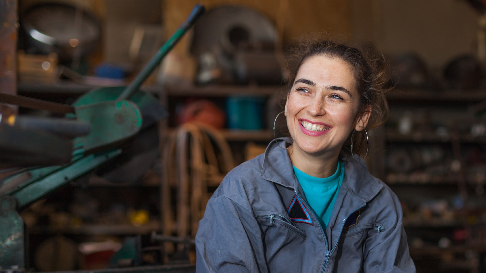 Junge Frau im Blaumann sitzt lächelnd in einer Werkstatt.
