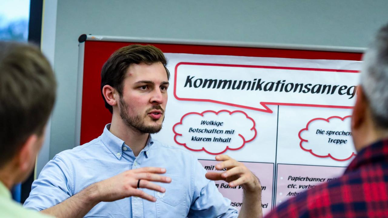 Ein junger Mann steht vor einer Tafel und gestikuliert.