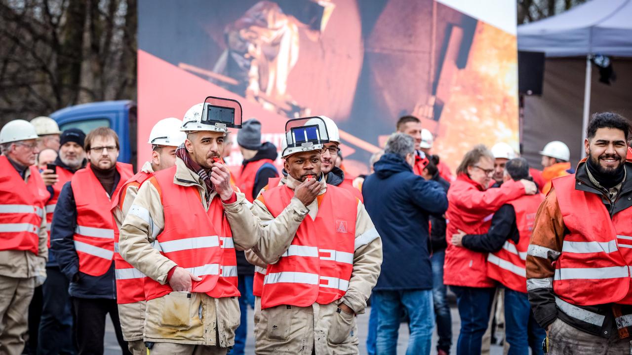 Zwei Männer mit Arbeitskleidung, Schutzhelmen und roten IG Metall-Westen blasen in Trillerpfeifen.