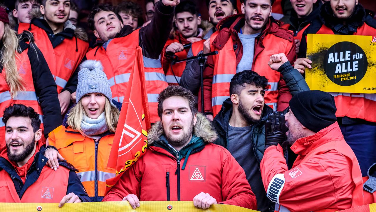 Zwei Männer halten ein Banner. Dahinter steh eine Frau mit einer IG Metall-Fahne. 