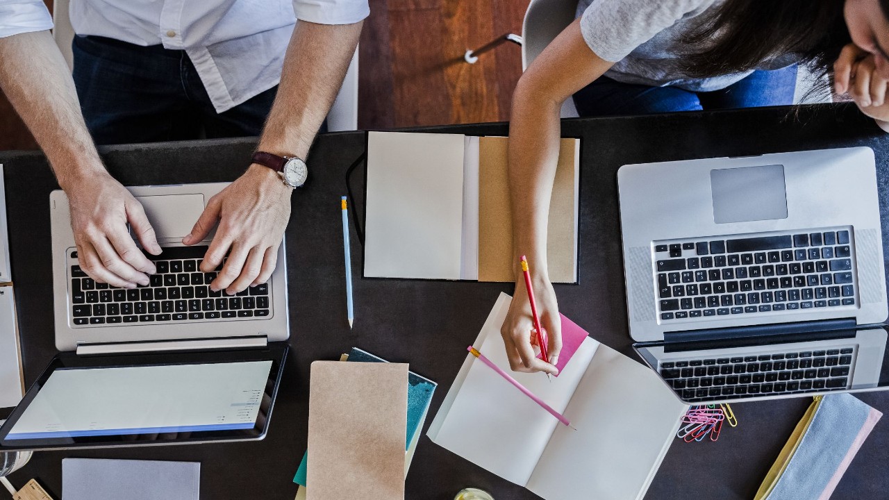 Zwei Personen arbeiten an Laptops und machen sich zeitgleich handschriftlich Notizen