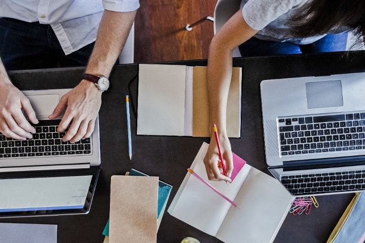 Zwei Personen arbeiten an Laptops und machen sich zeitgleich handschriftlich Notizen