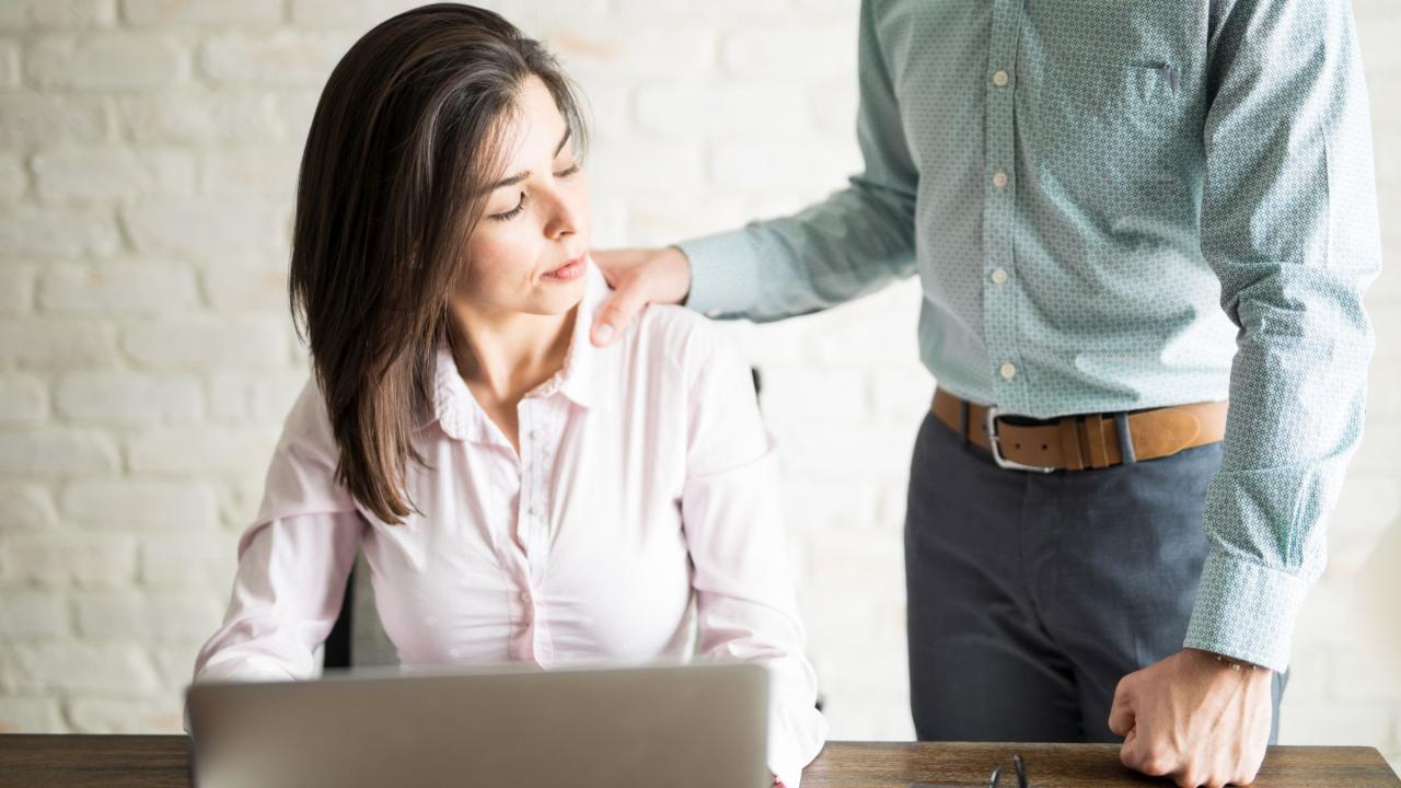 Mann fasst einer Frau an ihrem Arbeitsplatz von hinten an die Schulter