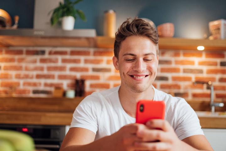 Ein junger Mann sitzt in der Küche und schaut in sein Handy.