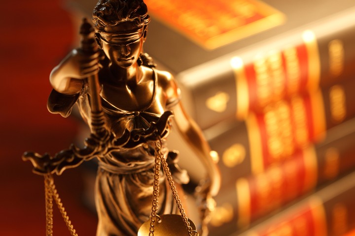 Justizia steht vor Bücherstapel mit Rechtstexten, sie hält eine Waage in der Hand und trägt eine Augenbinde