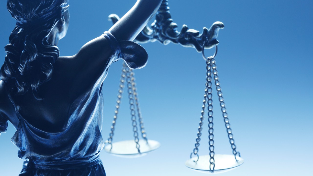 Ein Bild von Justizia als Bronzestatue vor blassblauem Hintergrund, auf dem sie eine Waage hält