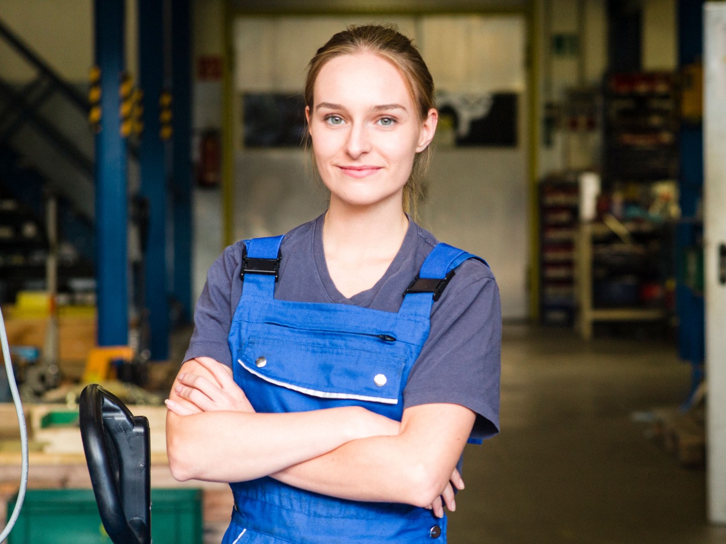 Eine junge Frau im Betrieb in einer blauen Arbeitshose und einem blauen T-Shirt steht mit verschränkten Armen da und schaut freundlich