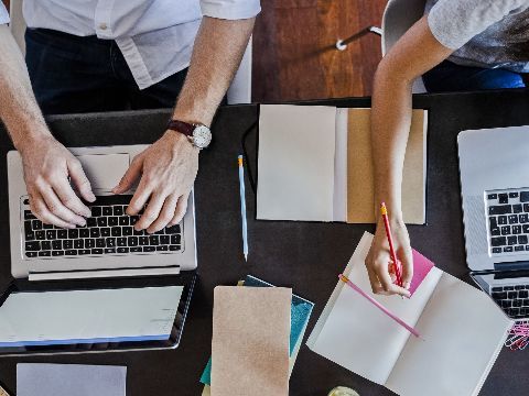 Zwei Personen arbeiten an Laptops und machen sich zeitgleich handschriftlich Notizen