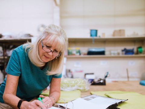 Eine Frau in einer Werkstatt beugt sich über einen Aktenordner und notiert etwas.