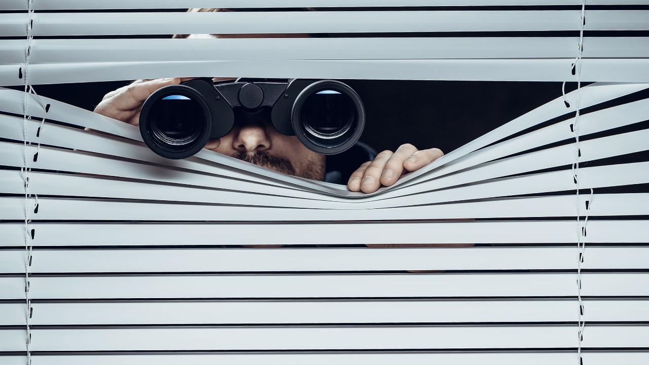 Ein Mann späht offenbar heimlich mit einem Fernglas durch eine Jalousie