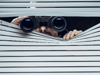 Ein Mann späht offenbar heimlich mit einem Fernglas durch eine Jalousie