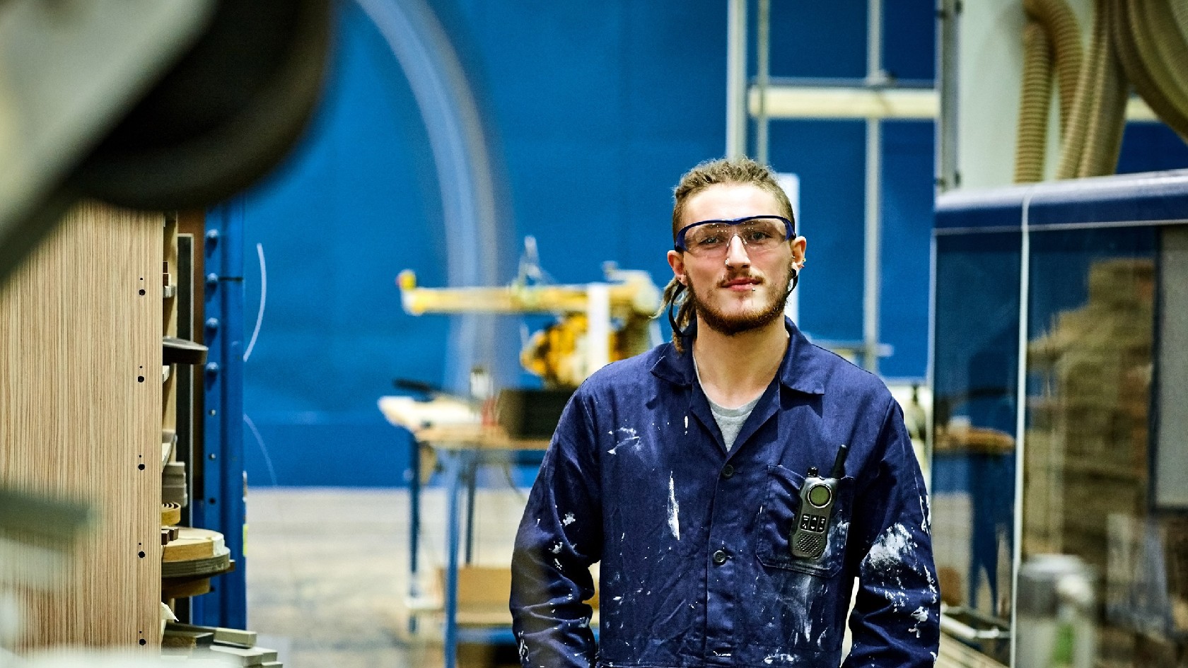Ein junger Mann im Blaumann mit Schutzbrille und Dreadlocks steht in einer Werkhalle und blickt lächelnd in die Kamera.