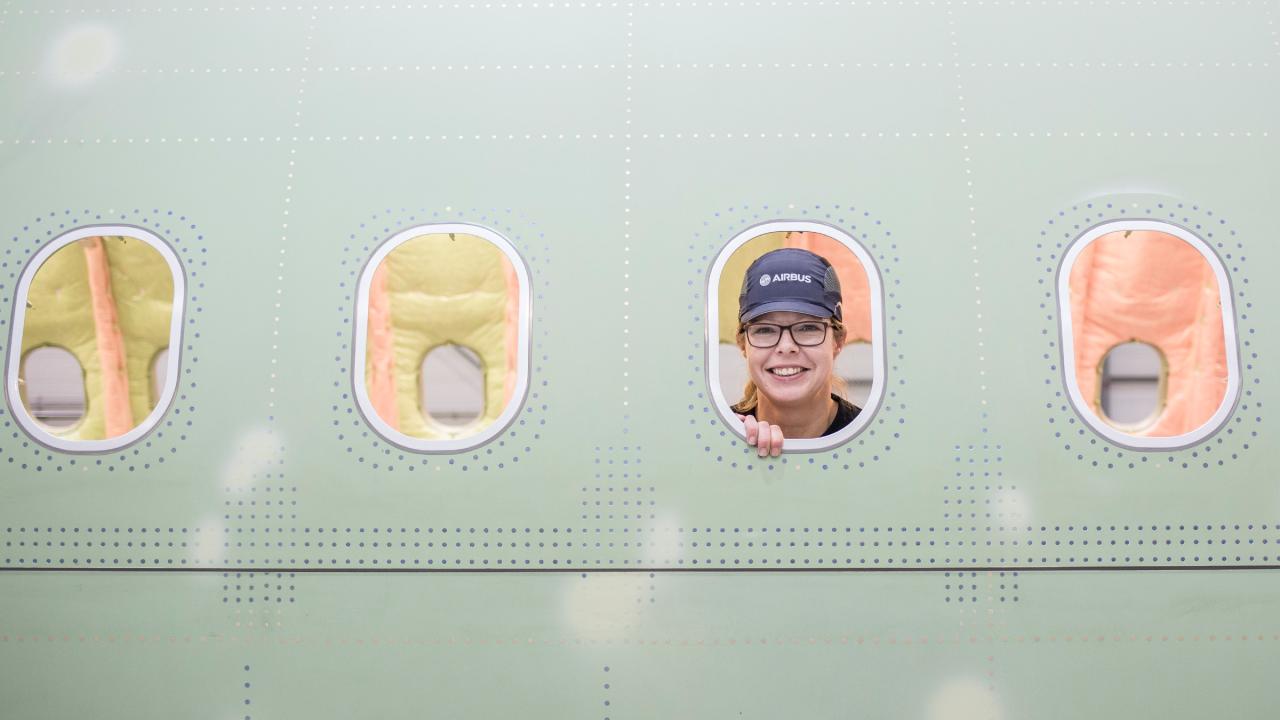 Eine Frau mit Kappe schaut durch das Fenster eines unfertigen Flugzeugs.